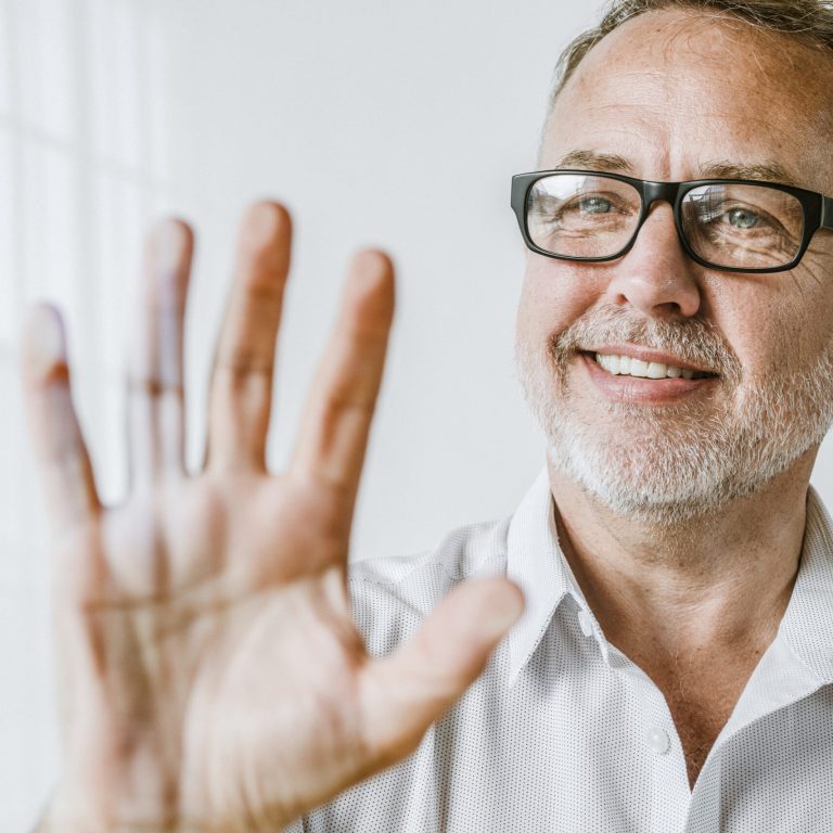 A smiling middle-aged man with glasses and a gray beard holds his hand up toward the camera, showing no visible Dupuytren’s contracture symptoms. He is wearing a light-colored shirt and standing in a bright, white room.