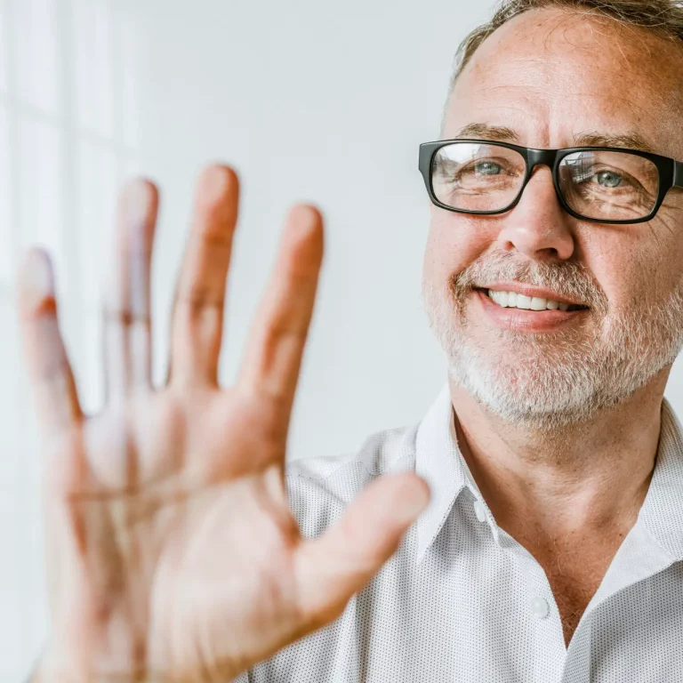 A smiling middle-aged man with glasses and a gray beard holds his hand up toward the camera, showing no visible Dupuytren’s contracture symptoms. He is wearing a light-colored shirt and standing in a bright, white room.