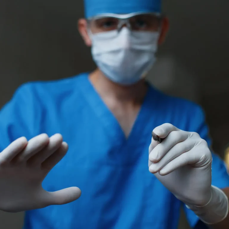 Young professional doctor in a blue medical uniform in rubber gloves in a white protective mask holds in his hand a metal scalpel in the operating room. Modern medicine and tools.