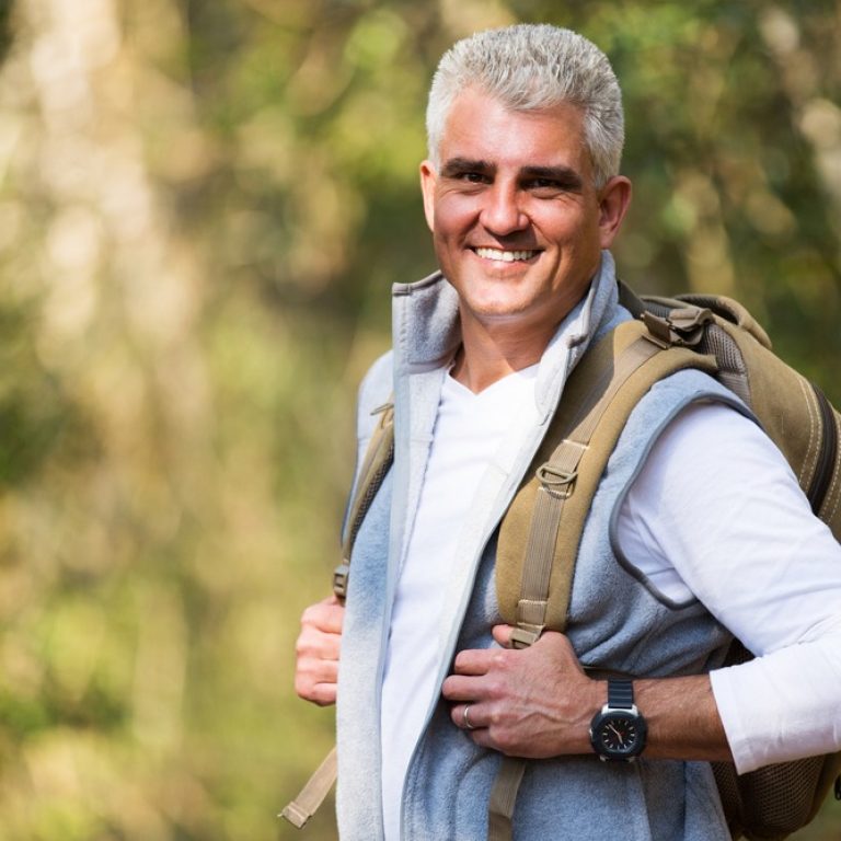 A smiling middle-aged man with short gray hair, wearing a white shirt, light gray vest, watch, and backpack, stands outdoors in a sunlit, wooded area—unaware of potential Dupuytren’s contracture symptoms that can affect hand mobility.